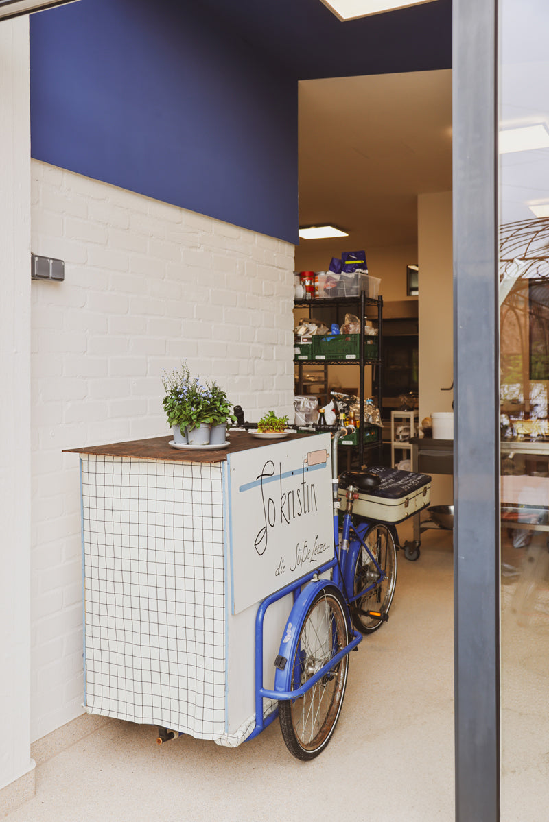 Im Eingang der Backstube der Patisserie steht ein umgebautes blaues Lastenrad. Vorne drauf steht eine weiße Vitrine mit Holzoberfläche auf der Blumen und ein Teller stehen. Die Backstubenwand ist weiß, und die Decke blau. Man sieht etwas weiter in die Backstube, ein Regal mit backzutaten, Metalltische und ein Bäckerei Ofen.
