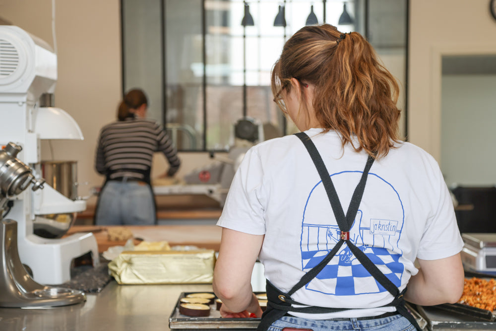 Jo Kristin steht in ihrer Backstube mit dem Rücken zum Betrachter. Man kann in die Backstube reinschauen und sieht hinter auch noch eine andere Mitarbeitern beim Arbeiten. Man schaut den beiden quasi beim Arbeiten über die Schulter. Es zeigt den Panoramablick, der Backstube.