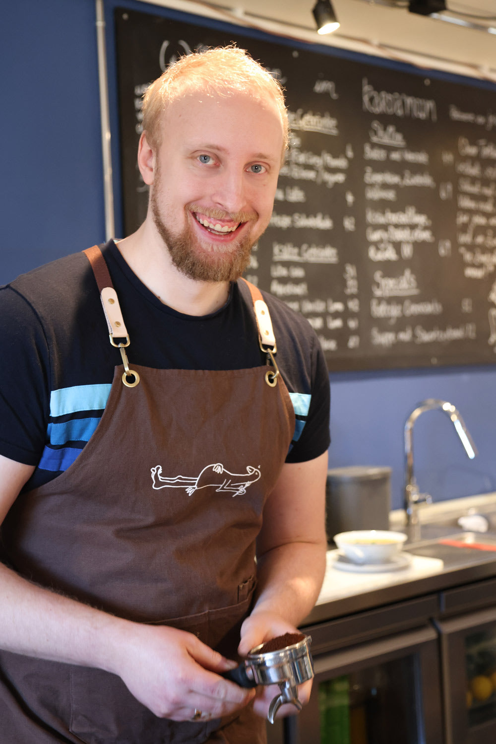 Max, der Koch und Barista im Cafe Kardamom steht hinter der Theke mit frisch gemalenem Kaffee in einer Gruppe einer Siebträgermaschine.