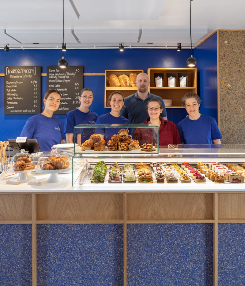 Im Vordergrund ist eine gefüllte Vitrine mit normalen und veganen Törtchen und Zimtschnecken sowie Croissants und herzhaften Stullen zu erkennen. Dahinter steht das sympathische Team mit Jo Kristin, der Inhaberin des Kardamom Cafés in Münster vor einer blauen Wand mit zwei Preistafeln und Regalen, die mit glutenfreiem Brot und Kaffee bestückt sind.