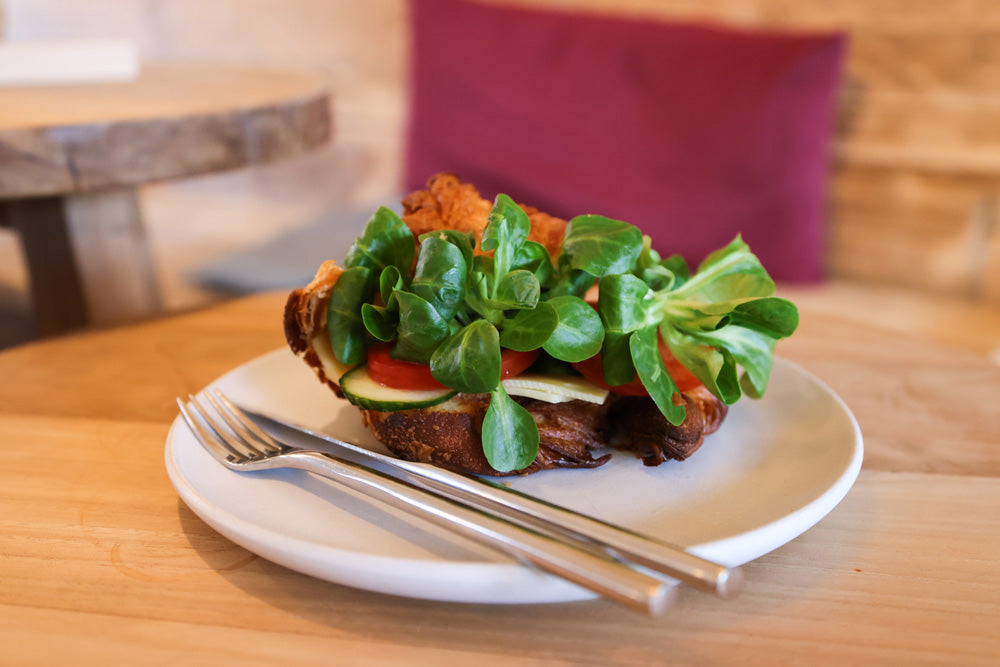 belegtes Croissant mit Käse, Feldsalat, Tomate und Gurke auf einem Teller liegend, auf einem Tisch im Kardamom in Münster stehend.