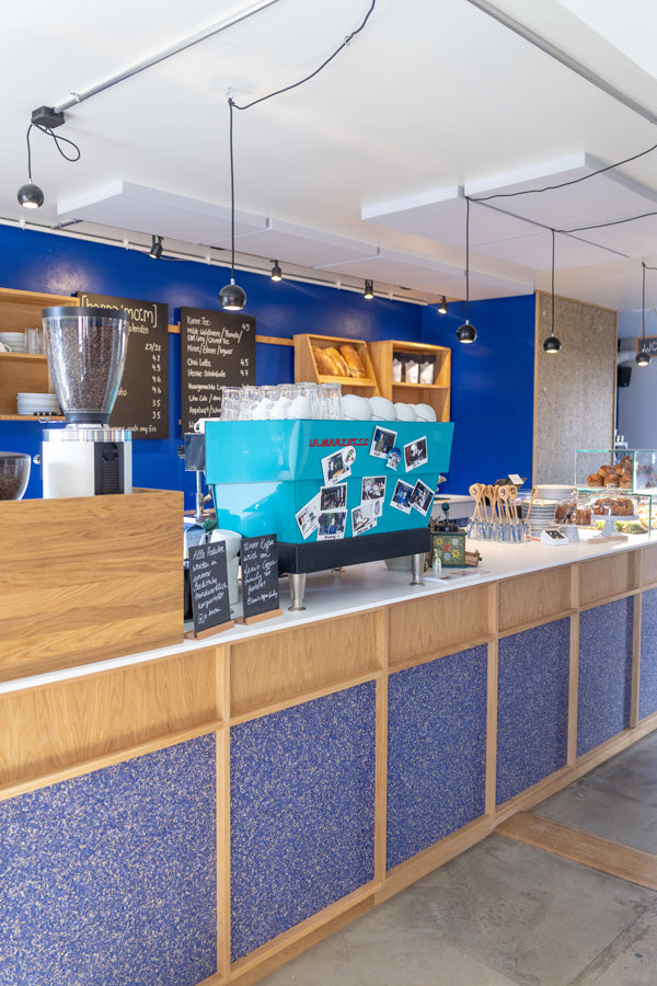 Kardamom Cafe Theke in Münster. Es steht eine türkisfarbene Kaffeemaschine auf einer Eichentheke mit Kornblumentapete und weißer Arbeitsplatte, wie auch Kuchenvitrine vor einer blauen Wand. An der Wand hängen Eichenregale und Tafeln mit Getränkekarte.