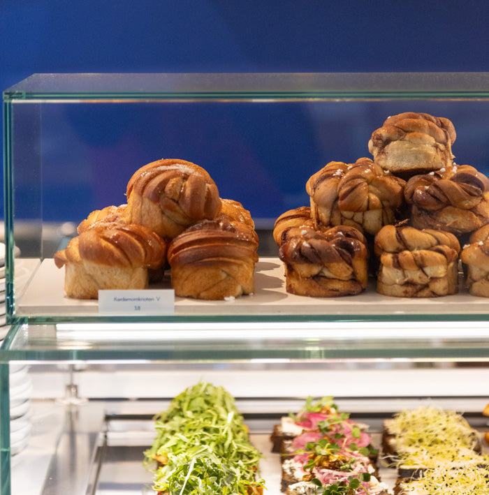 Kardamom und Zimtschnecken in der Vitrine im Kardamom Cafe. Im Hintergrund ist eine blaue Wand und in der unteren Etage sind belegte Stullen zu sehen.